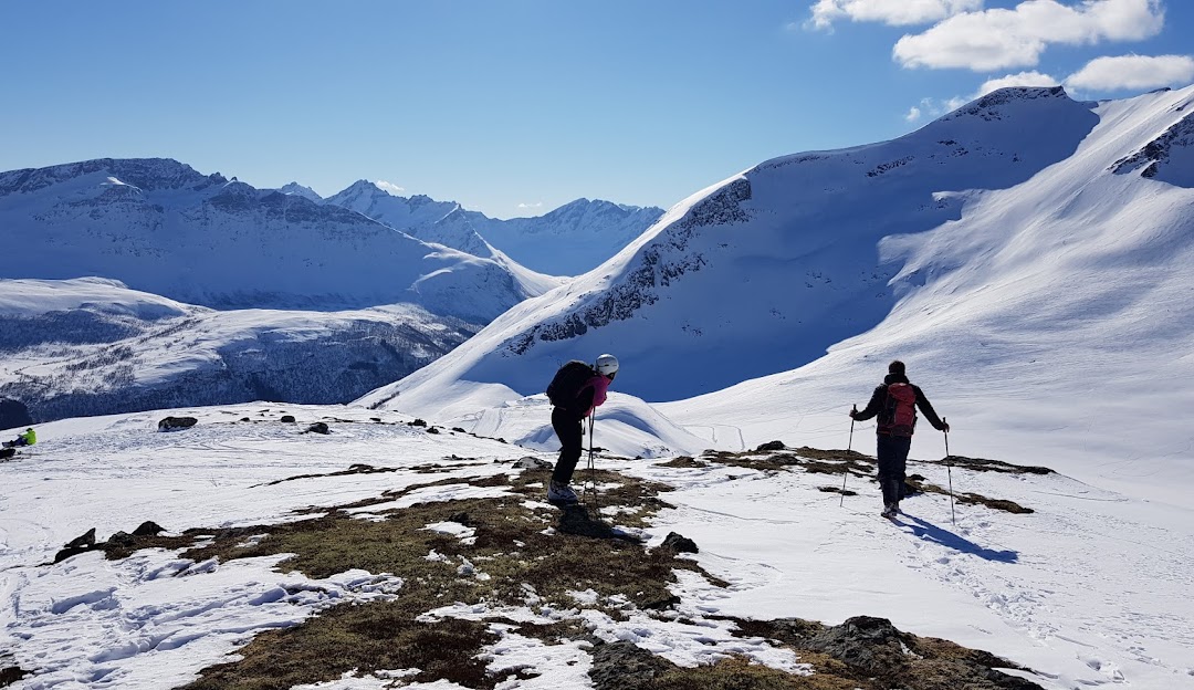 Winter sports scene at Sunnmørsalpane Skiarena Fjellsætra in Straumgjerde, Norway, featuring a chalet with a stunning backdrop of snow-covered mountains and a skier enjoying the slopes.