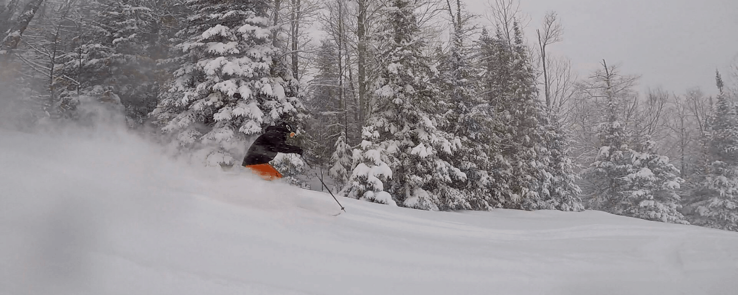 A skier skillfully navigates a snowy slope at Ski Brule in Iron River, Michigan, with a snowmobile parked nearby. The ambiance of a lively winter sports scene is discernible, featuring distant figures possibly skiing or snowboarding and a faint ski lift.