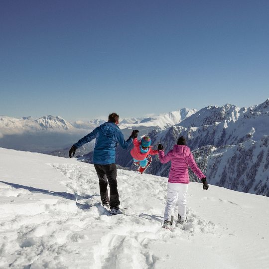 A family enjoying a skiing session in Hochzeiger – Jerzens in Tyrol Austria. The winter sports scene is complemented by a challet and a wider view of the ski resort.