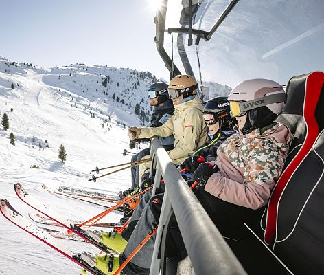 A family enjoying a day of skiing at the Hochzeiger - Jerzens resort in Tyrol, Austria, with a ski lift and various winter sports activities in the backdrop.