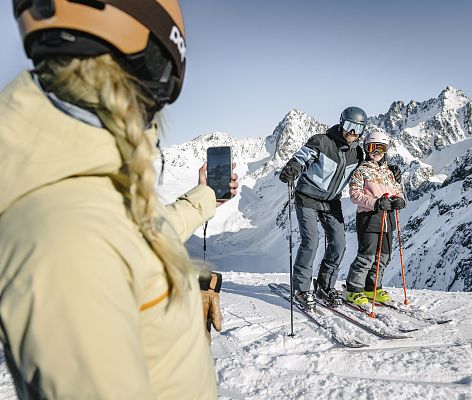 A winter scene at Hochzeiger–Jerzens Tyrol Austria featuring a family skiing down the lush snow-capped mountains a ski lift running in the background and a picturesque challet on the side.