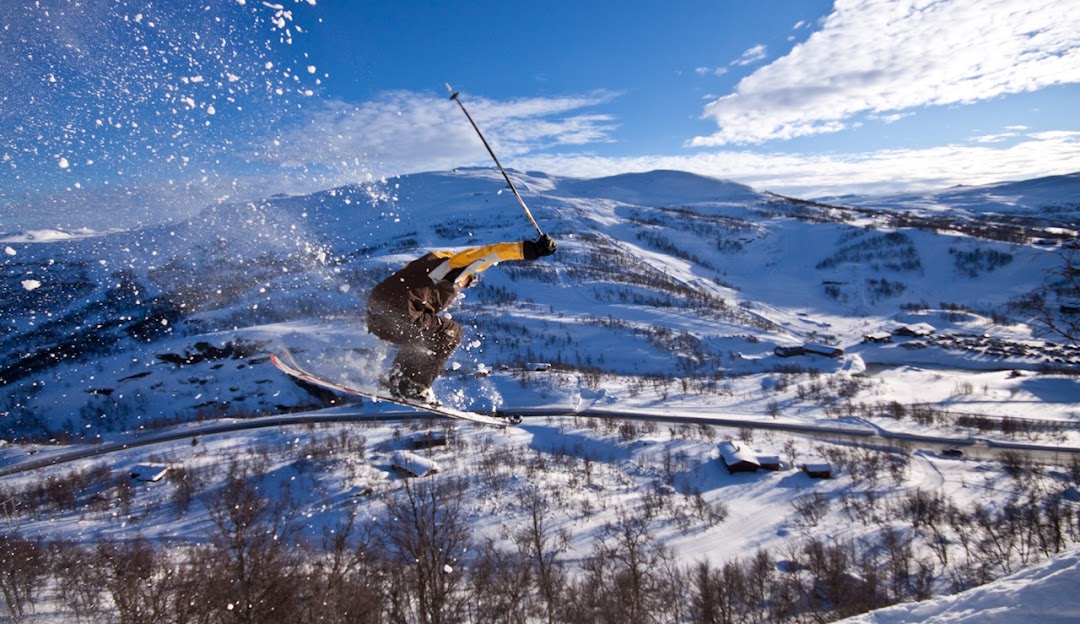 A skier gliding down the snowy slopes in Tyin-Filefjell, Norway, amidst a scenic winter sports scene. Visible elements include a distant ski lift and vague figures snowboarding.