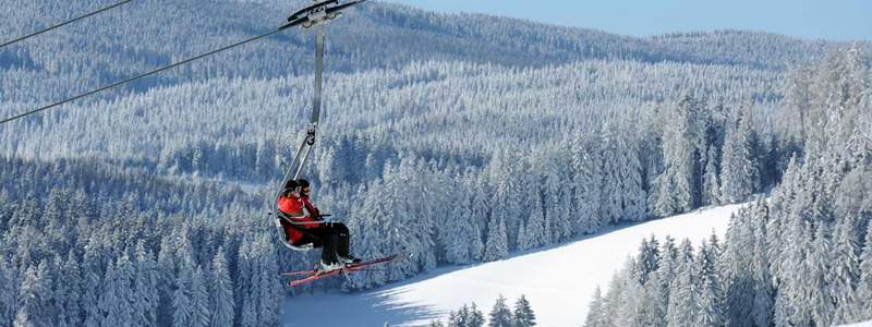 Winter scene at Ski-Waldheimat Hauereck resort in Styria, Austria featuring a ski lift, skiers in action and a charming chalet nestled in the snowy landscape.