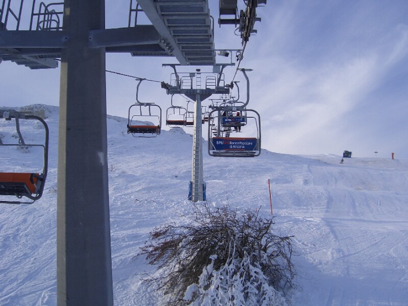 Skiers enjoying downhill skiing at Risalita sciistica Carpegna-Eremo, Italy with a ski lift in the foreground and a picturesque chalet nestled amongst snowy trees.