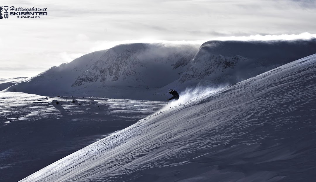 A skier enjoying a winter sports scene in Hallingskarvet, Norway. The landscape also features a majestic mountain in the background and a snowmobile nearby.