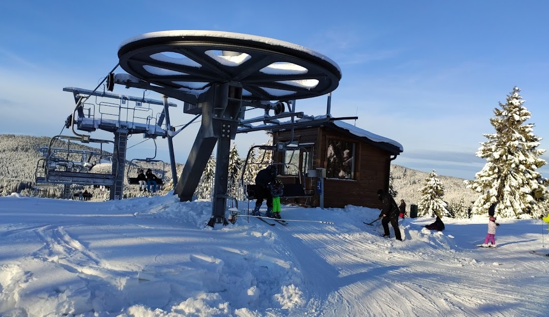 A view of Orava Snow in Slovakia, featuring a ski lift ascending a snowy hillside amidst a ski resort. Winter sports enthusiasts dot the landscape. A cozy challet peeks from the background.