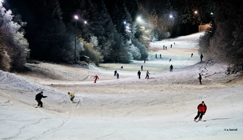 A vibrant winter scene at Alpe del Nevegal, Italy, showcasing a busy ski resort with numerous skiers enjoying the slopes, a ski lift operating in the backdrop and a cozy chalet nestled in the powdery snow.