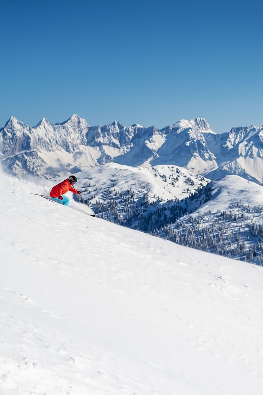 A skier enjoys the snow at Zauchensee | Flachauwinkl ski resort in Austria, surrounded by a typical winter sports scene, including a chalet and a distant snowboarder.