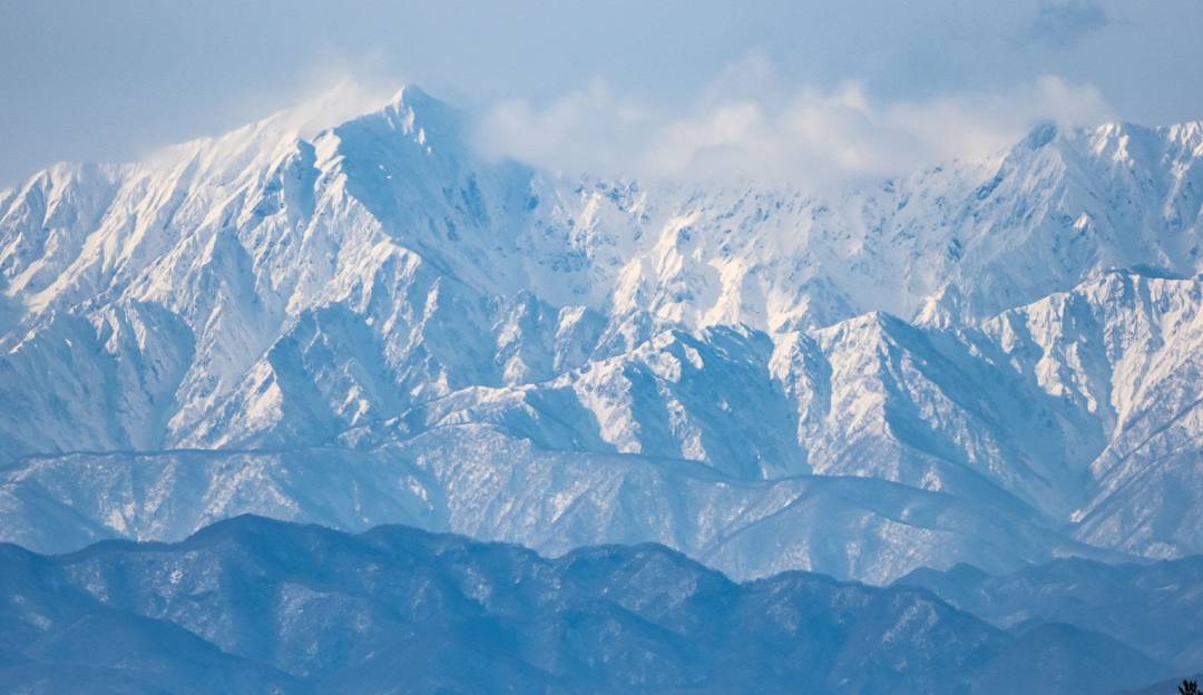 View of a towering mountain covered in snow at Shigakogen Hasuike in Nagano, Japan. A quaint chalet is nestled at its base amidst a stunning winter scenery with sparse visible winter sports activity.
