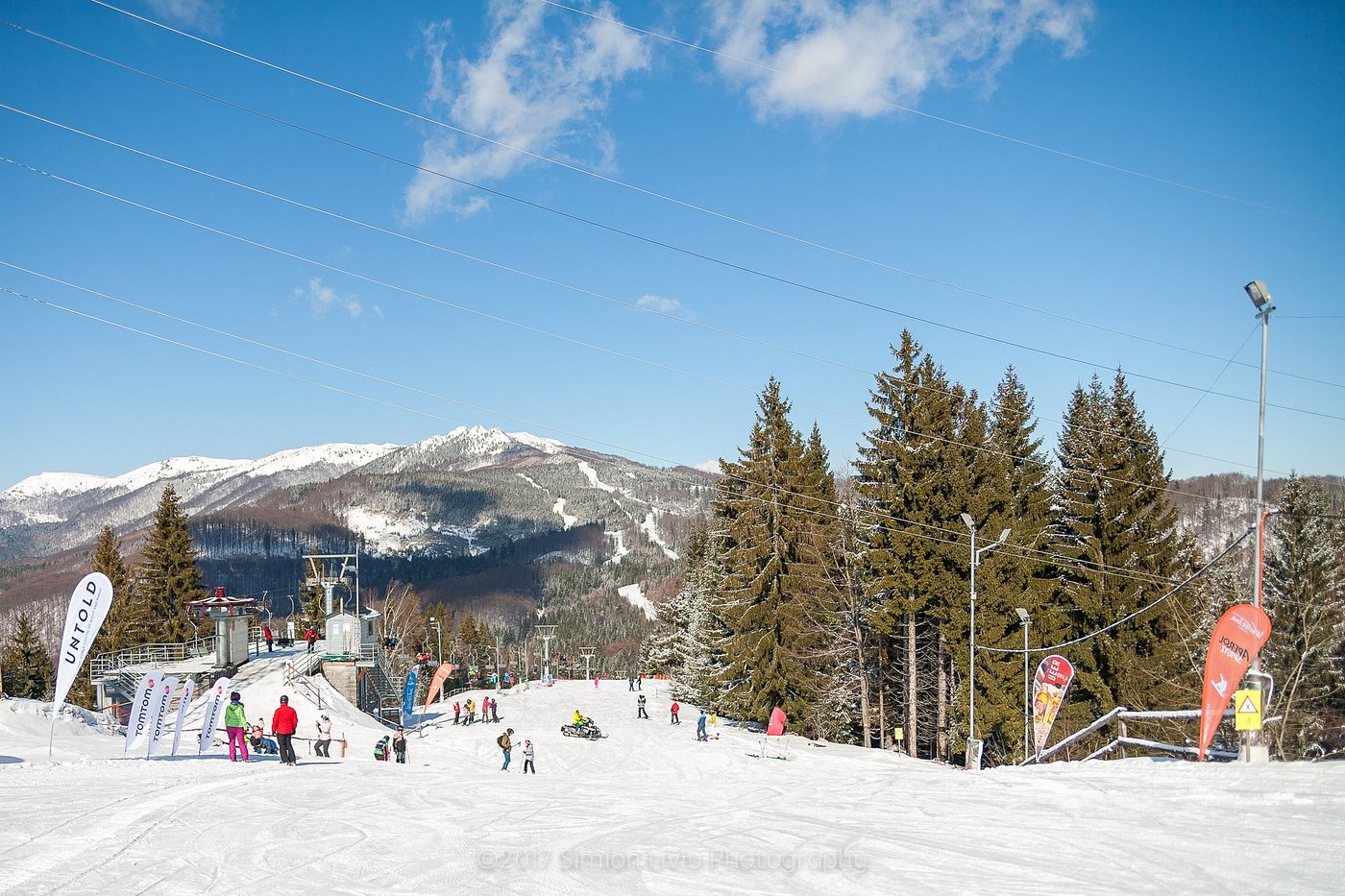A lively winter scene at Superski Roata Cavnic in Romania featuring a ski resort nestled amidst snow-covered slopes. Skiers maneuver down the hills with a ski lift seen in the background.