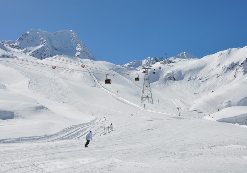 Ski resort at Stubai Glacier in Austria, showing a lively winter sports scene. Skiers, a chalet, and a winter sports centre are among the features visible in the snowy landscape.