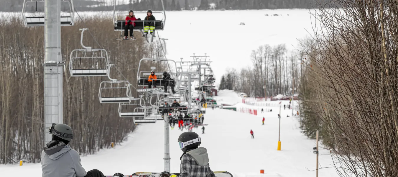 Winter sports scene at Kinosoo Ridge in Cold Lake, Alberta, featuring a ski lift ascending the mountain, busy with skiers at the ski resort. A snowmobile is discernible in the distance.