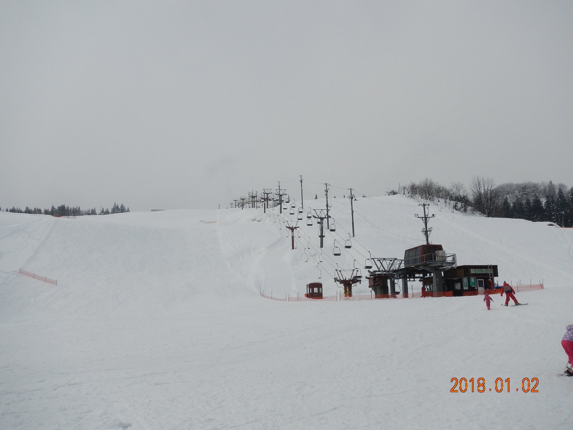 Winter scene at a ski resort in Itsukamachi, Japan, featuring a ski lift ascending snow-covered slopes towards a cozy hilltop challet.