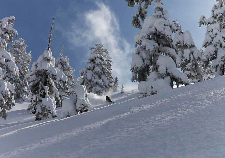 A skier gliding down a snowy slope at Mount Cain Alpine Park, a ski resort on Vancouver Island, with the towering mountain backdrop and a ski lift nearby.