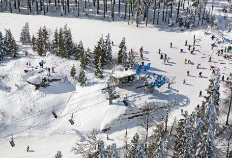 The image features a lively winter sports scene at Špičák ski resort in Czech Republic with skiers riding the ski lift and a chalet in the background amidst a beautiful snowy landscape.