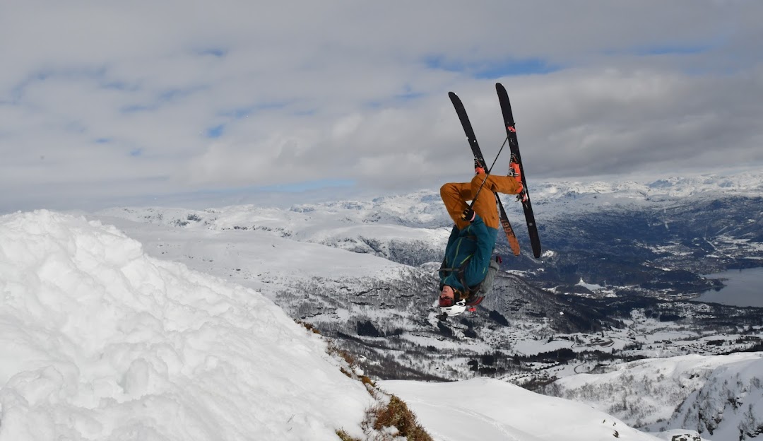 A skier confidently glides downhill at Sauda Skisenter in Western Norway, a picturesque winter sports scene unfolding in the background. Nearby, a snowboarder takes to the slopes while a quaint chalet and operational ski lift are subtly visible.
