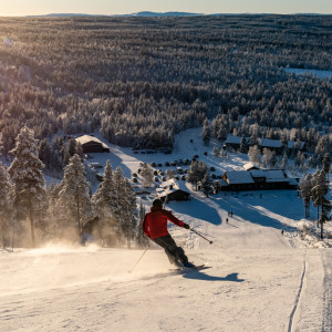 A skier and a snowboarder are enjoying a beautiful winter day at the Tännäskröket ski resort in Northern Sweden. They are moving down the snow-covered slopes with a ski lift visible in the background.