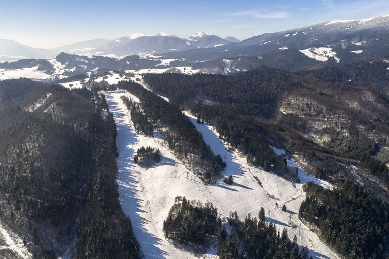 A winter scene at Ski Park Racibor in Slovakia featuring a ski resort with a chalet and people enjoying winter sports. A ski lift can barely be seen in the background.