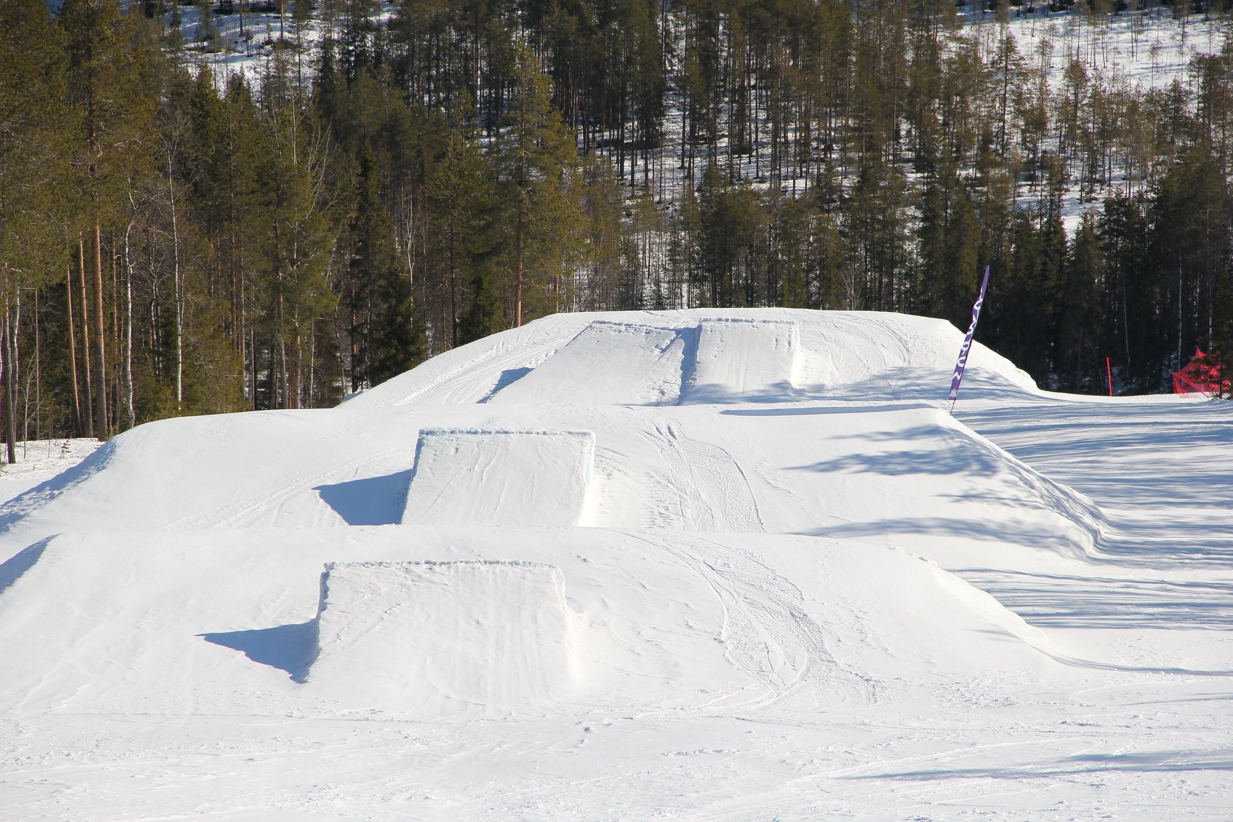 A skier enjoys a winter sports scene at Suomutunturi in Lapland, Northern Finland, with glimpses of a snowboarder, ski resort, and a snowmobile in the distance.