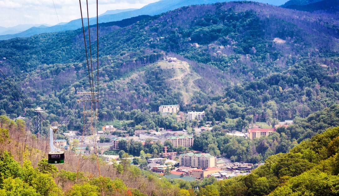 A scenic view of Ober Mountain in Gatlinburg Tennessee showcasing a charming chalet amidst verdant landscapes. A ski lift is visible hinting at the ski resort nearby all illuminated under a bright sunny day.