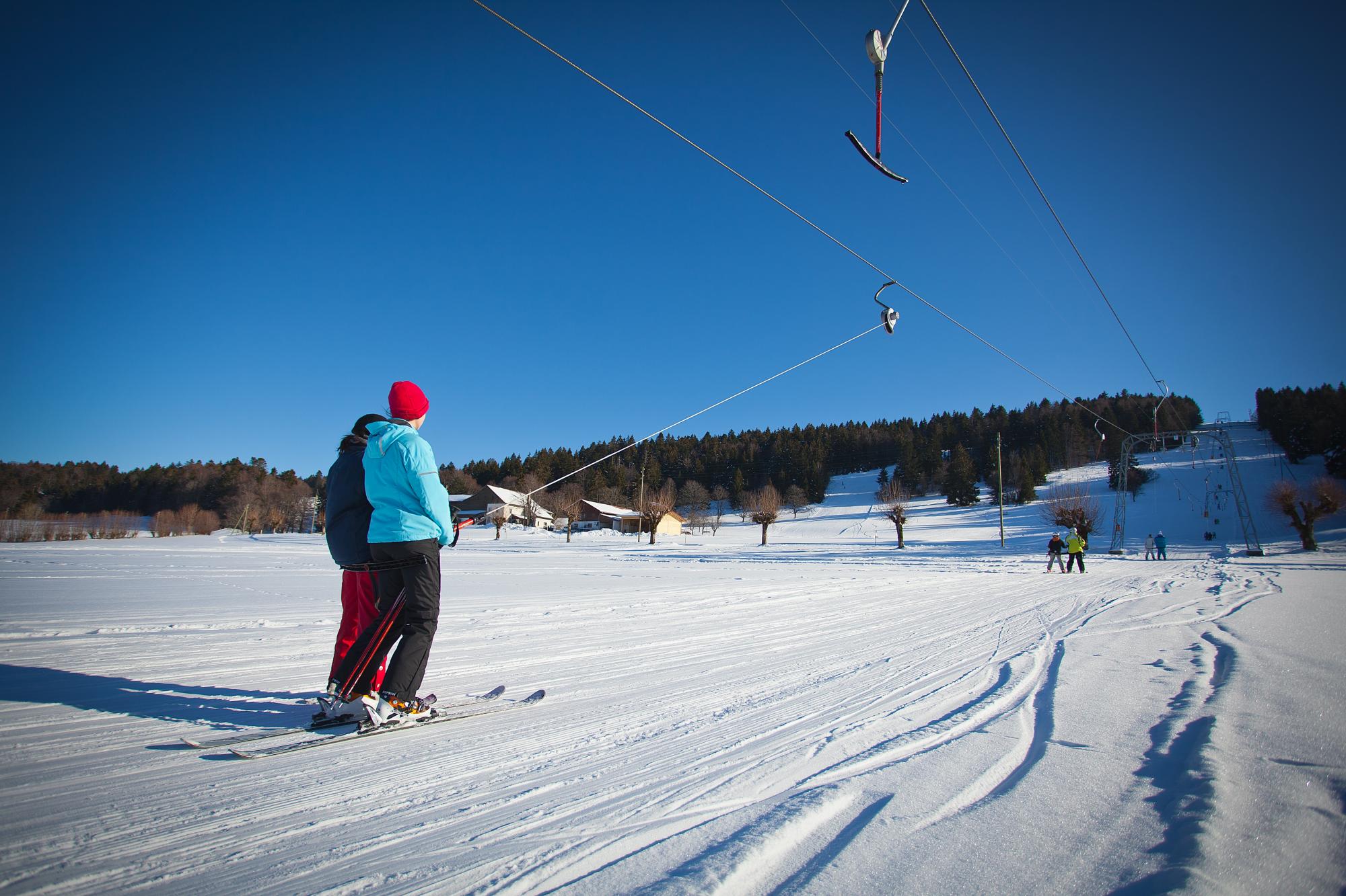 A skier enjoying a winter sports scene in Tramelan, Switzerland, with a child learning to ski nearby. A ski lift is visible at the winter sports centre in the background.
