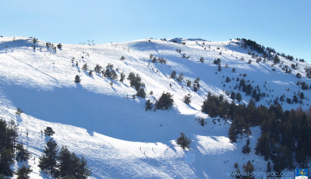 Ski resort in Camurac, France featuring snow-covered slopes bustling with winter sports enthusiasts. A charming chalet overlooks the scene and a single skier can be seen in the distance.