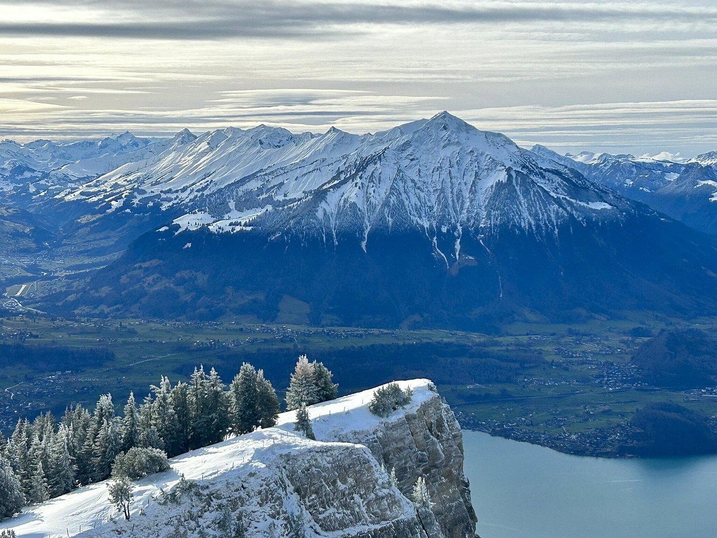 A captivating view of Beatenberg-Niederhorn in Switzerland showcasing a large mountain covered in snow. Winter sports enthusiasts can be seen enjoying the scene. A charming chalet sits subtly in the frame all enveloped by serene winter scenery.
