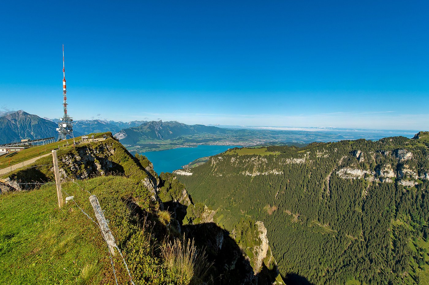 Scenic view of a sunny day in Beatenberg-Niederhorn, Switzerland, showcasing a charming chalet nestled against towering mountains, the calm lake in proximity, and a mountain bike implying a day of adventure.