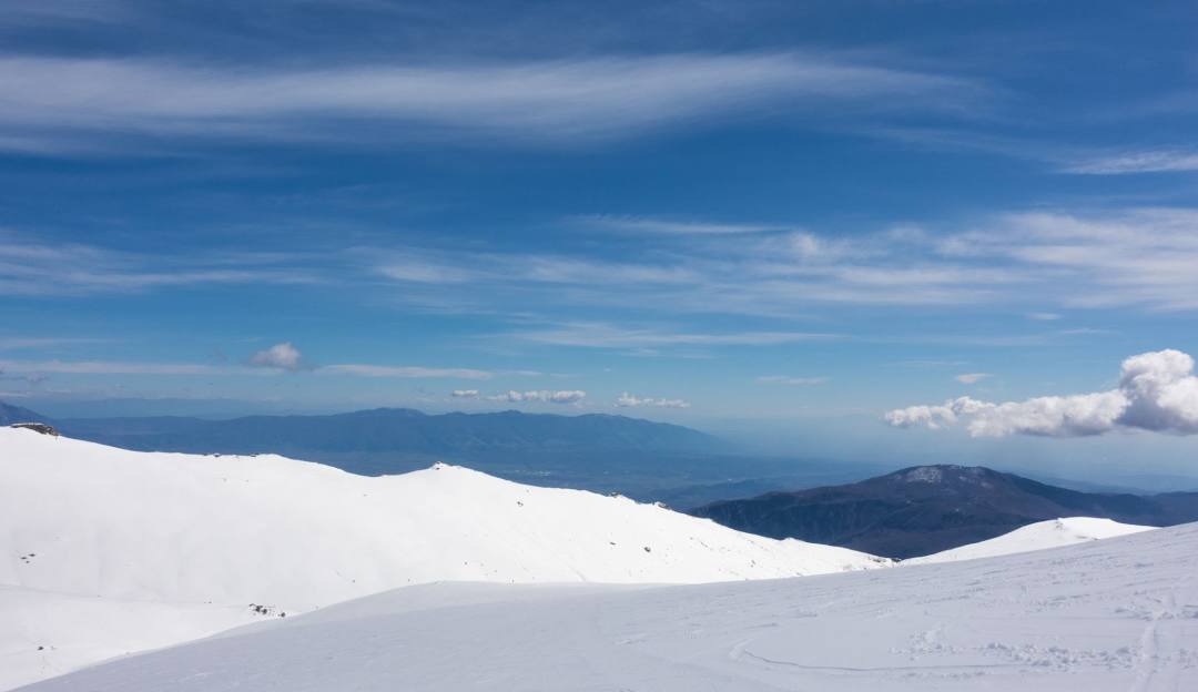 A skier traverses down Kaimaktsalan in Edessa, Greece. A charming challet sits in the distance as a snowy mountainous backdrop paints a picturesque winter sports scene.