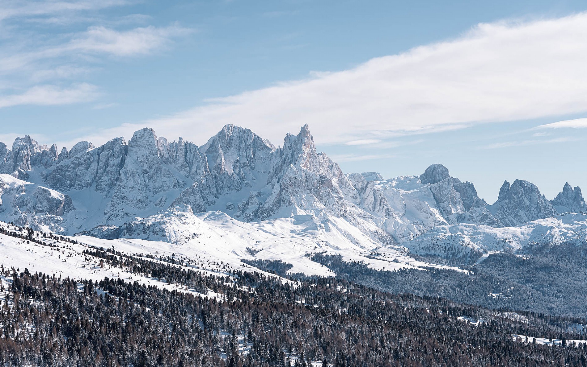 Scenic winter view of Alpe Lusia in Trentino Italy featuring a majestic snow-covered mountain a quaint challet in the foreground and distant figures engaging in winter sports activities.