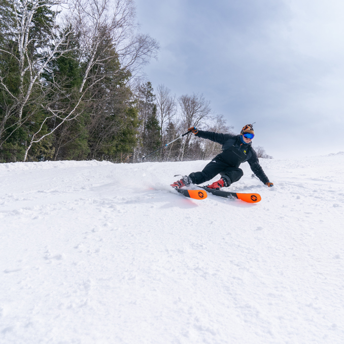 A winter sports scene at Sommet Gabriel in Quebec Canada featuring a skier and a snowboarder on powdery slopes with snowmobile and ski resort vaguely visible.