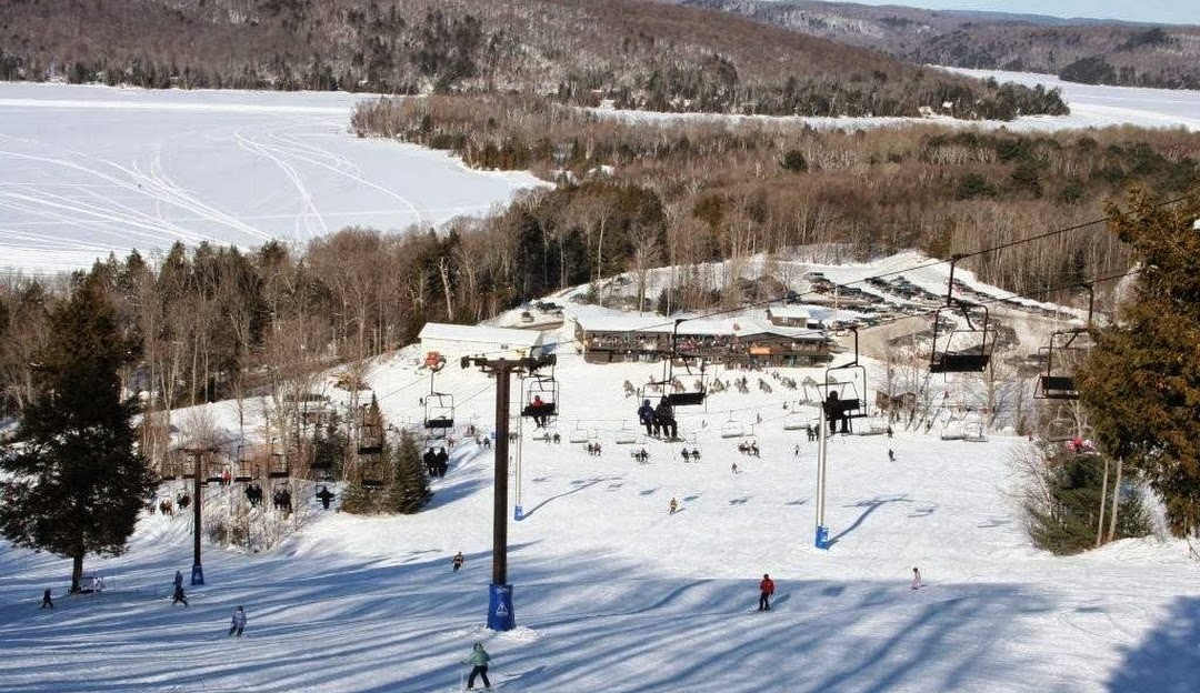 View of Sir Sam's Ski Resort in Eagle Lake, Ontario, with people enjoying various winter sports. A ski lift, a challet, and snow blanketing the scene round out this Canadian winter tableau.