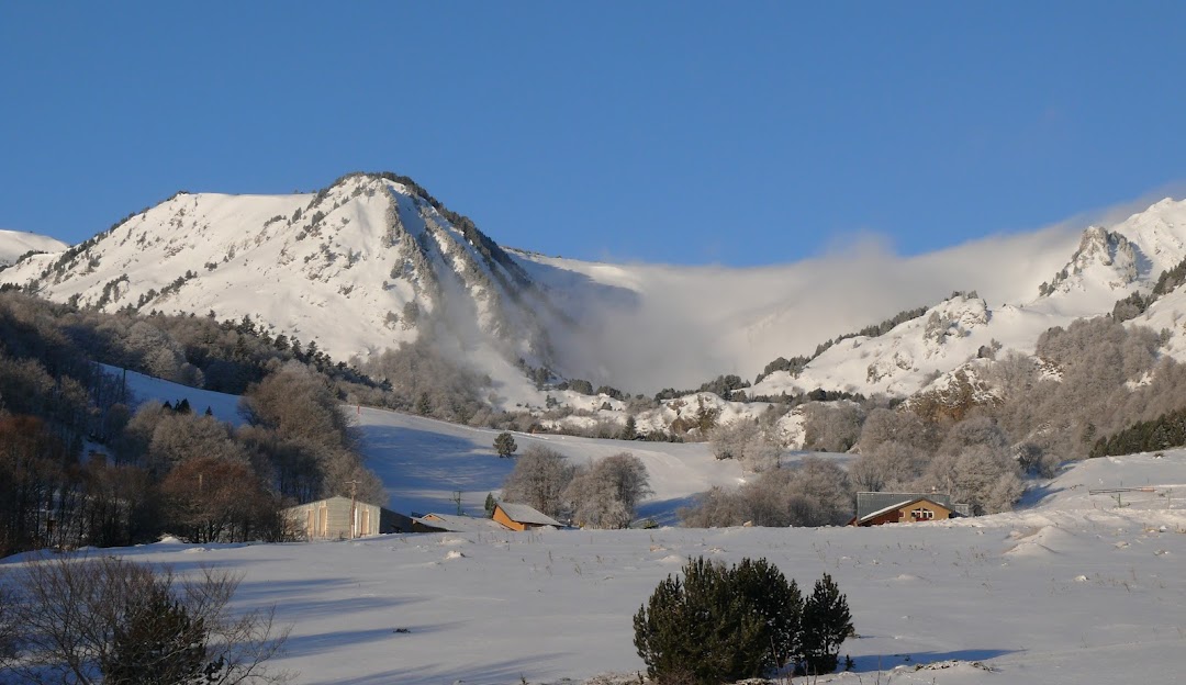 Image of a charming chalet nestled in the snowy winter landscape of Mijanès-Donezan in Ariège, Foix, France. A winter sports centre and mountain are faintly visible in the background.