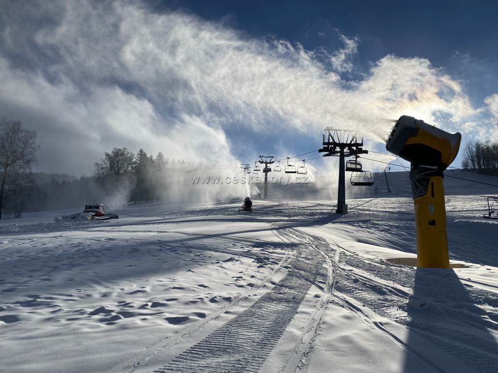 Winter sports scene at Deštné ski resort in Czech Republic featuring a ski lift a skier and a snowmobile among snowy slopes and coniferous trees.