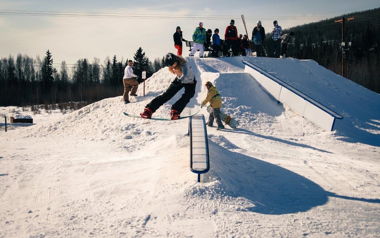 A snowboarder carves a path down the slopes at Birch Hill Ski & Snowboard Area in Fort Wainwright, Alaska, surrounded by pristine snow and clear blue skies.
