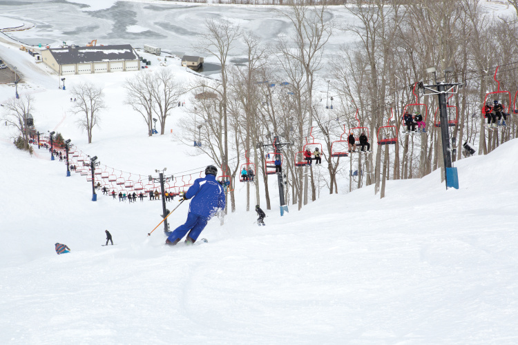 A winter sports scene at Perfect North Slopes in Lawrenceburg Indiana showcasing a skier gliding down the snowy slopes with beautiful winter scenery in the background. A snowboarder can be spotted at a distance.