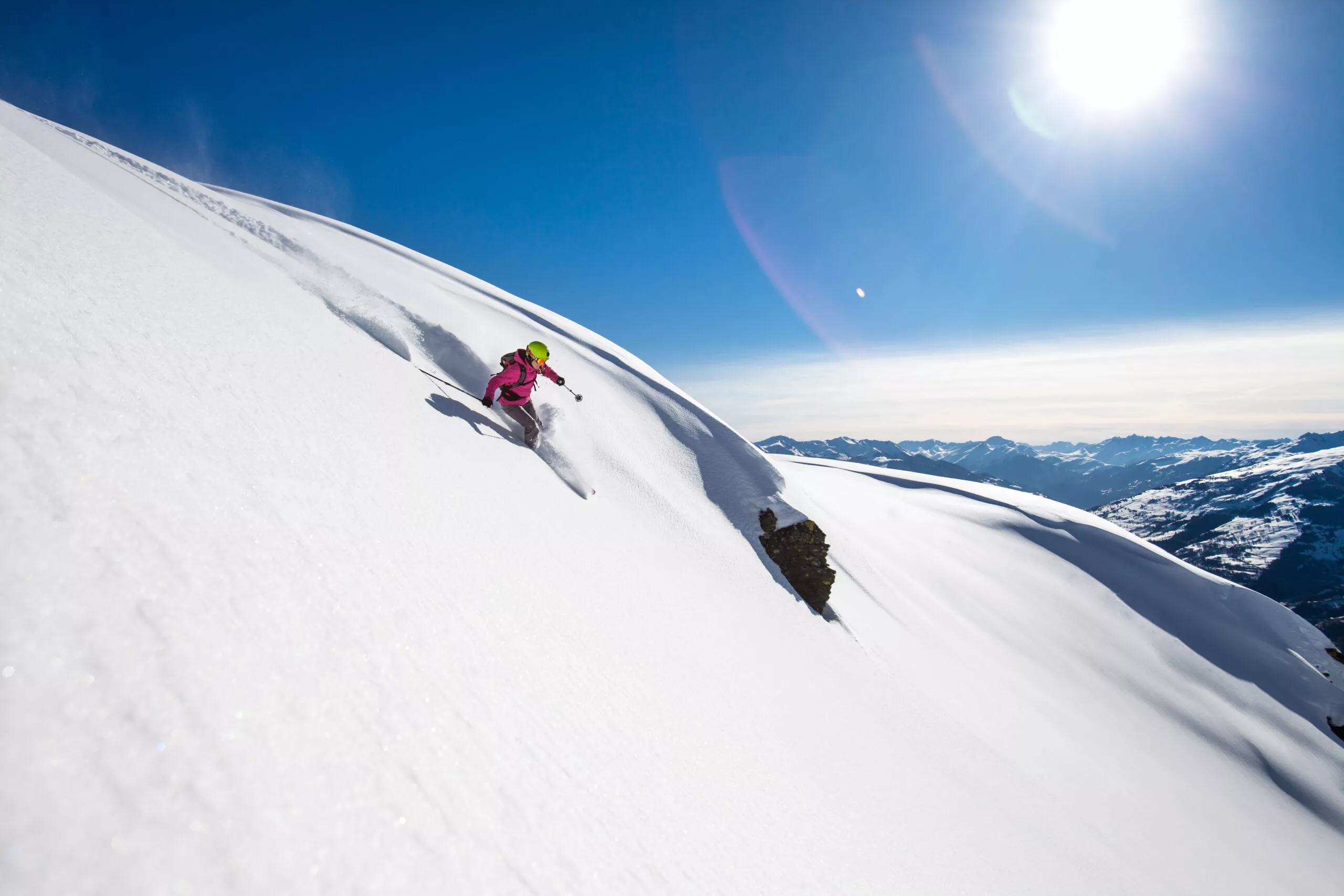 A skier navigating a snowy landscape in the Ruine Super Saint-Bernard area in Switzerland with a chalet in the distance. Small groups of people can be seen in the background participating in winter sports.