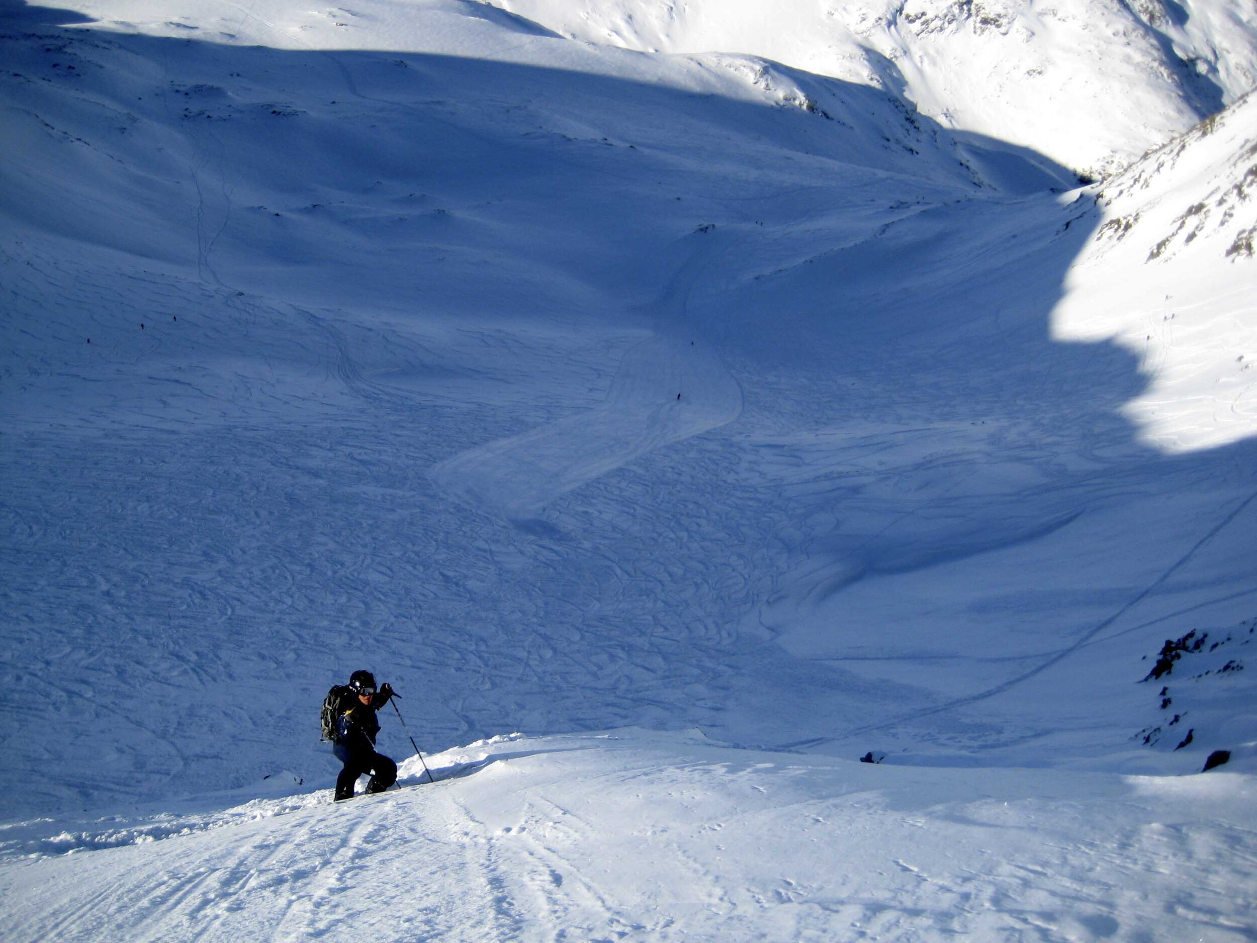 A skier swiftly glides down a wintery slope near the Super Saint-Bernard ruins in Valais Switzerland. Nearby a chalet peeks out from the snowy landscape. In the distance a snowboarder trails behind.