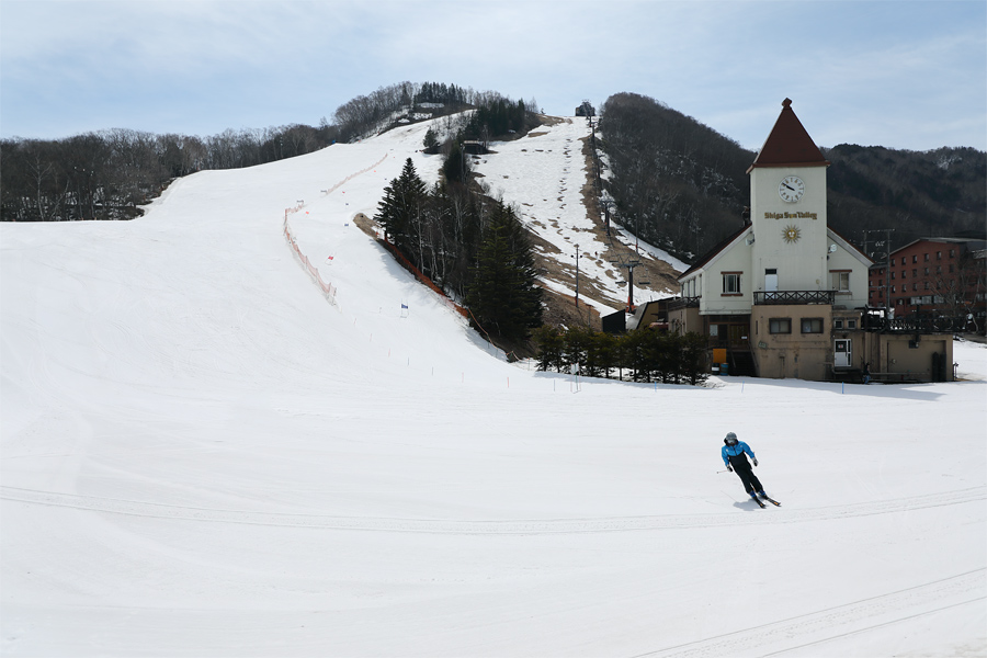 A skier navigating snowy slopes at Hoppo-Bunadaira Ski Area in Yamanochi, Japan. The image features a winter sports scene with a ski resort and lift visible in the background.