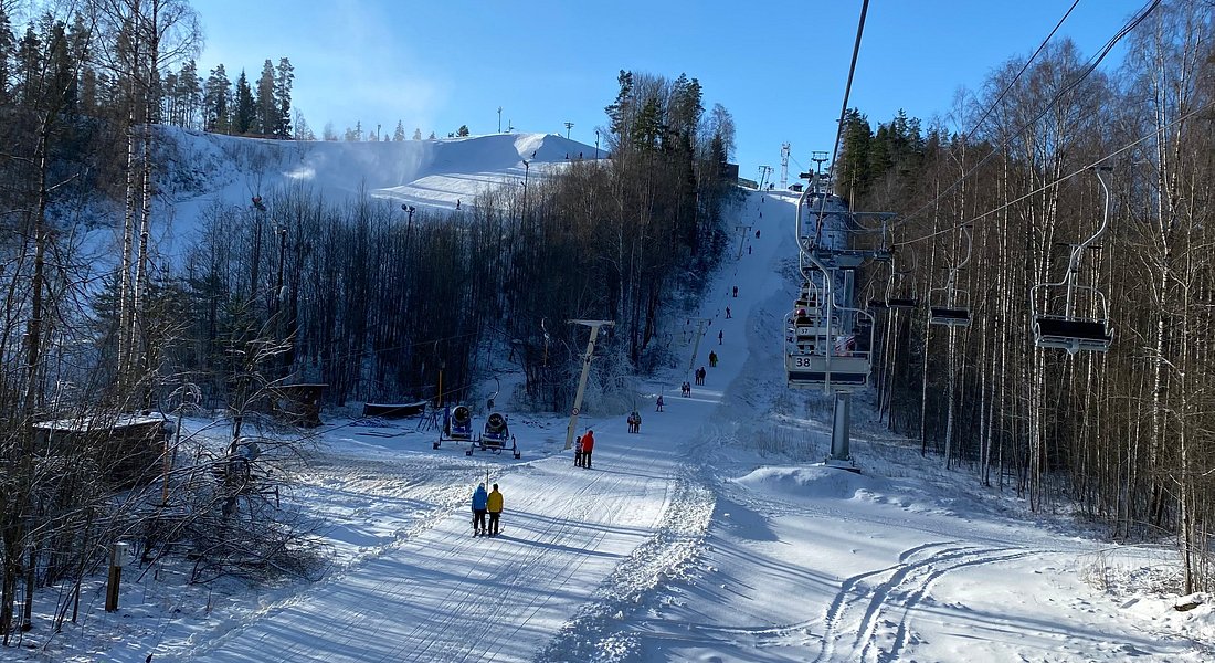 Ski resort scene at Krasnoye Ozero in Korobitsyno, Russia, featuring a bustling winter sports activity, visible ski lift, and a skier descending the slope.