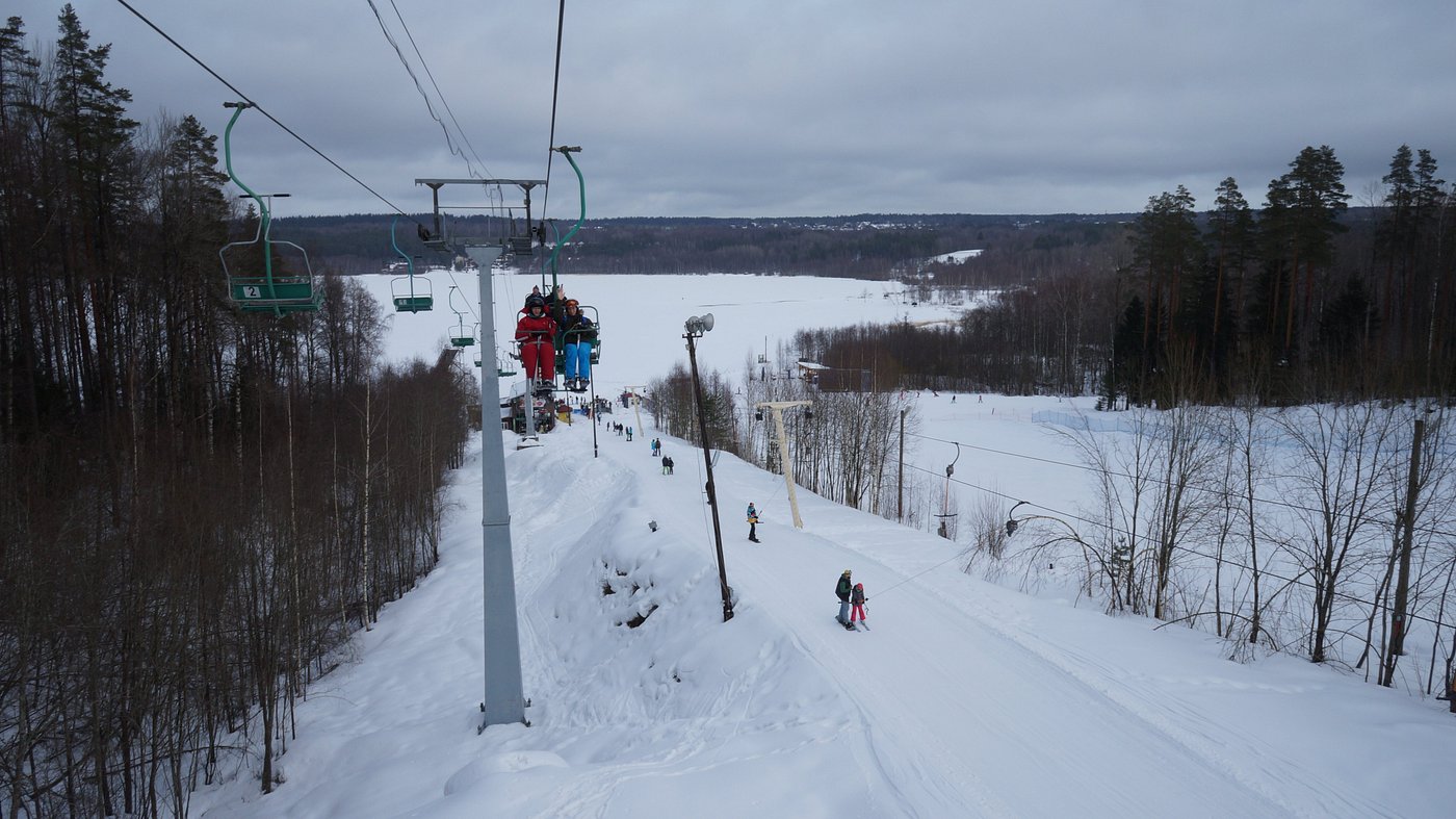 A winter sports scene at Krasnoye Ozero, Russia, featuring a ski lift transporting skiers up the hills while a skier proficiently glides down a hill, and a chalet visible in the backdrop.