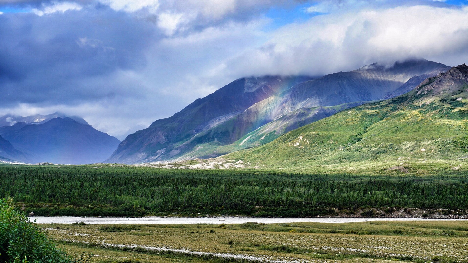 View of a majestic mountain peak under a clear sky at Black Rapid, Alaska. A chalet is nestled against the foothills, amid the serene landscape. A lake can be seen in the distance.