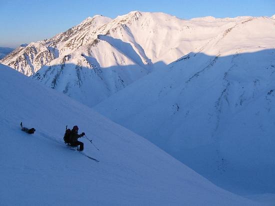 A skier swiftly maneuvers down a slope at Black Rapid in Alaska, USA, under clear winter skies. A cozy chalet waits invitingly in the backdrop, exhibiting the lively atmosphere of this charming ski resort.