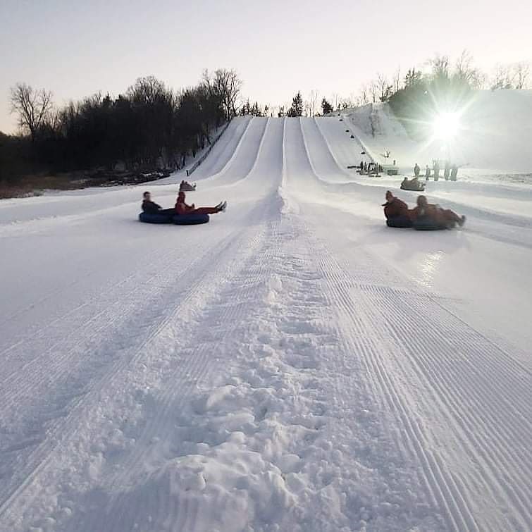 A lively winter sports scene at Seven Oaks, Iowa, featuring a snowmobile moving across a pristine white snowy landscape, with families and groups engaging in skiing activities.