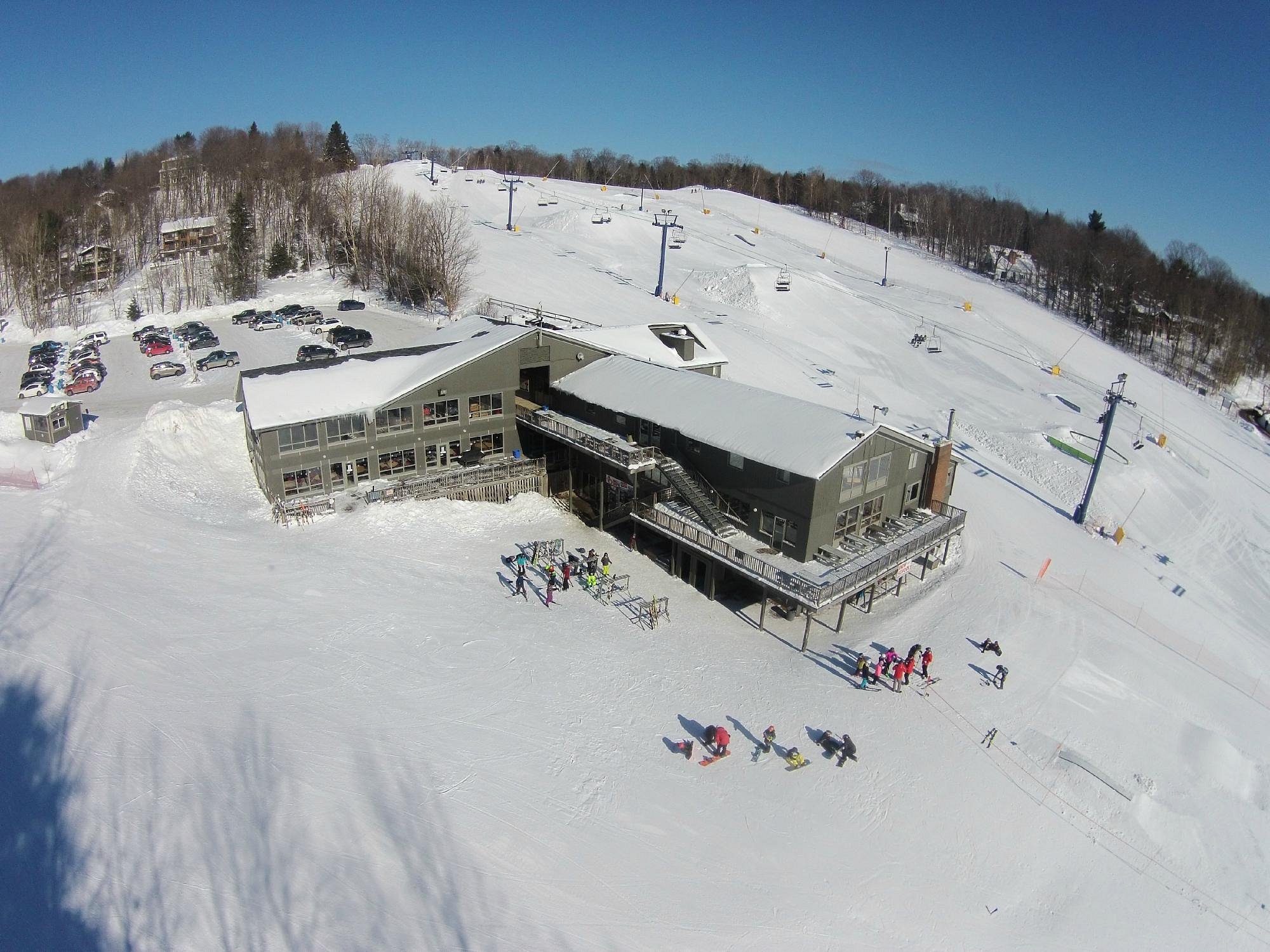 A woodland winter scene in Hidden Valley Highlands, Northern Ontario, featuring a bustling ski resort with skiers on the slope and an operational ski lift. Nestled among the trees, a charming chalet is also visible.