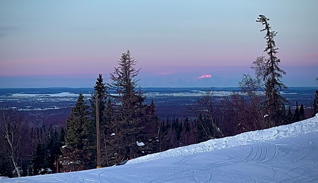 Winter scene at Hilltop Ski Resort in Anchorage, Alaska. A skier glides down a snowy hill, while a ski lift ascends, and a quaint chalet nestles in the background.