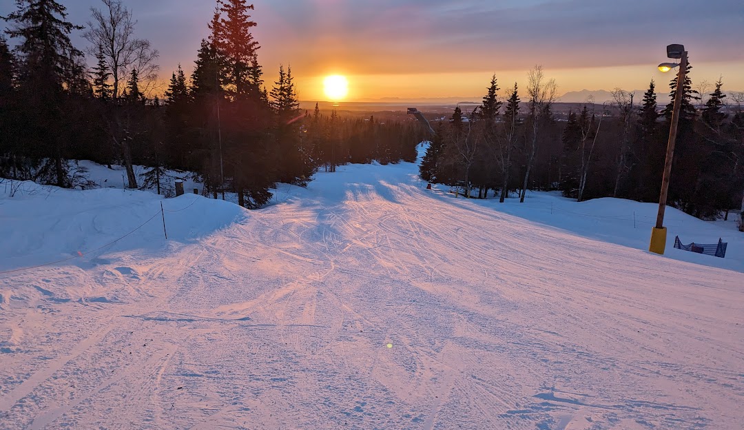 A winter sports scene at Hilltop – Anchorage in Alaska featuring a skier descending a snowy slope at a ski resort with a charming chalet and picturesque winter scenery in the background.