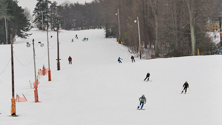 A bustling winter sports scene at Victor Constant Ski Resort in Hudson Valley New York with a skier in motion others on the ski lift and groups of people enjoying the snowy slopes.