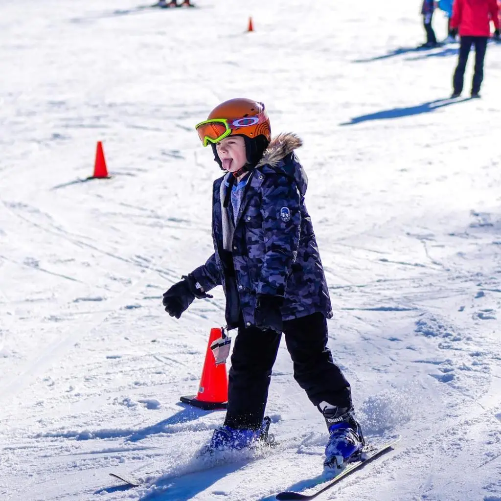 A skier and a child learning to ski dominate a lively winter sports scene in Little Switzerland, Wisconsin, with a hint of a snowboarder and chalet in the background.