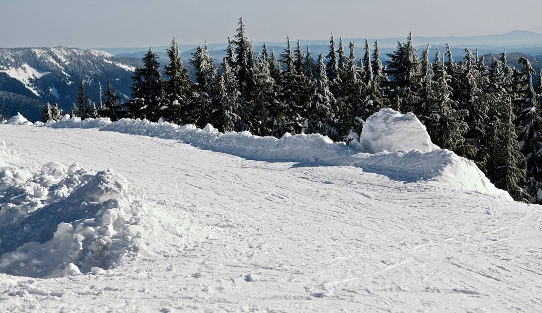 A winter sports scene at Timberline Summit Pass, Oregon with a skier and snowboarder navigating the snowy slopes. A snowmobile is parked nearby, and a chalet peeks out in the background.